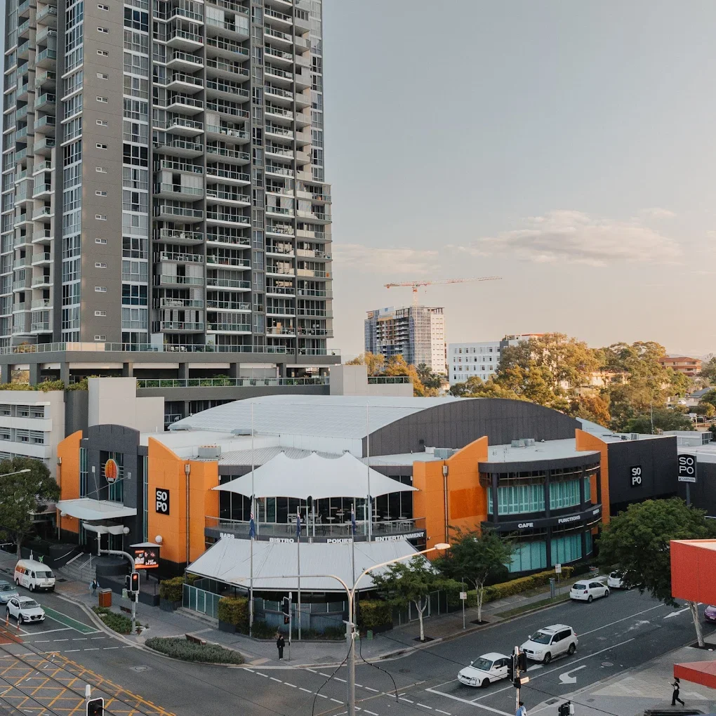 A city street view showing a multi-story building with balconies, a shopping center with orange and black exterior walls, a white canopy, and several cars and pedestrians on the street.