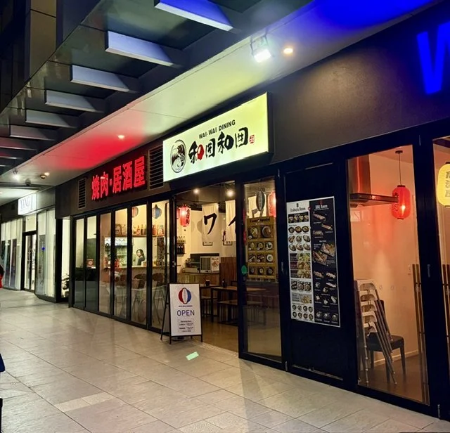 Night-time outside view of Japanese restaurant 'Wai-Wai Dining' in Southport showing glass frontage lit up by multiple colourful signs and lanterns inside.