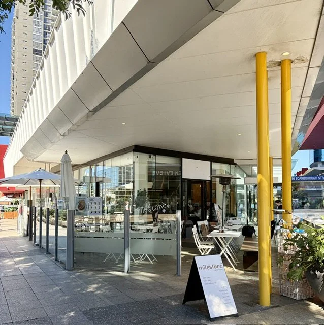 Street view of 'Milestone Cafe' in Southport showing the modern glass front with glass fenced seating area, tables and chairs, sun umbrella's and some greenery.