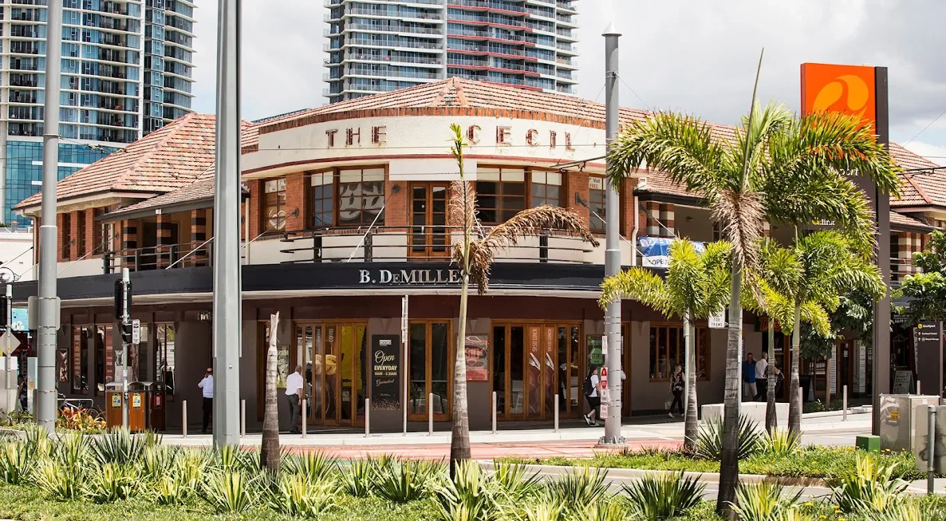 The Cecil Hotel. A two-story commercial building with a brown tiled roof, palm trees in front, and people walking by on the sidewalk.