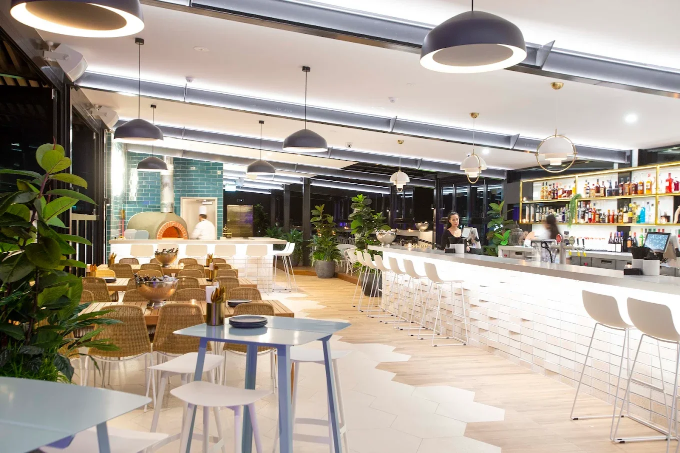 The Aviary, a modern, rooftop bar and restaurant in Southport with interior white counter, white bar stools, green plants, and shelves with bottles and glasses.