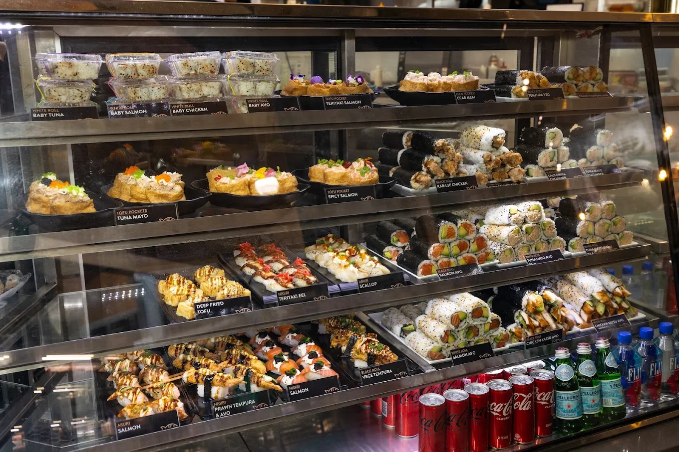 Assorted sushi rolls and sashimi in a glass display case with bottles of soda and water below.
