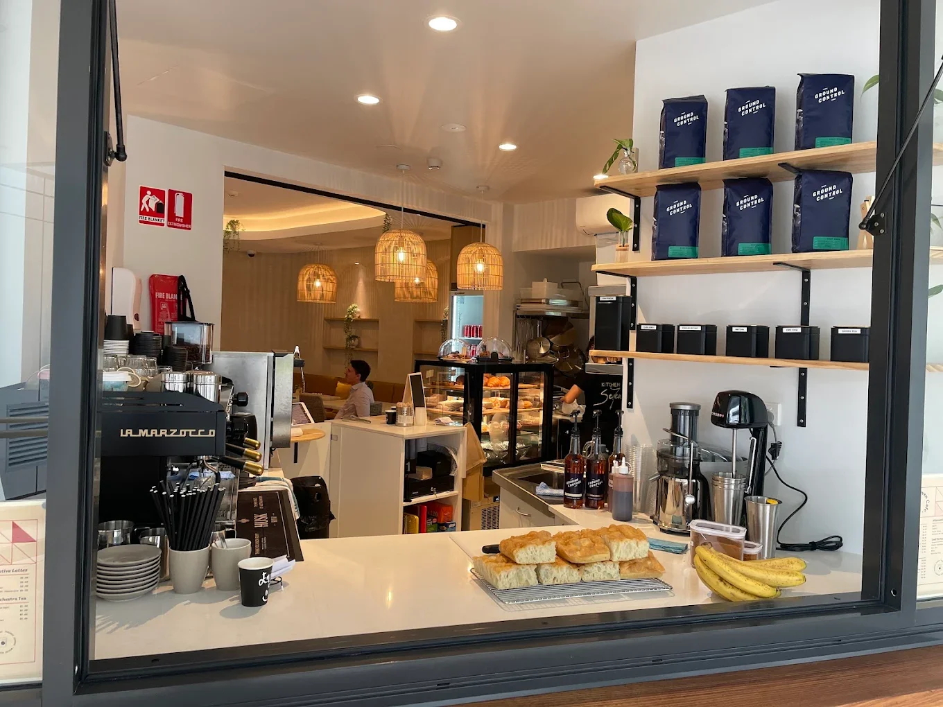 View of a cafe counter with baked goods, bananas, coffee-making equipment, shelves with coffee beans, and a person sitting in the background.