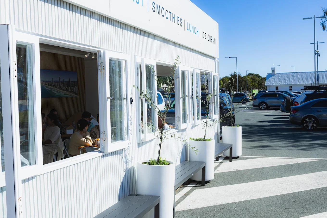 Outside of a white building with open windows, restaurants or cafe interior visible, parking lot with cars, and clear blue sky.