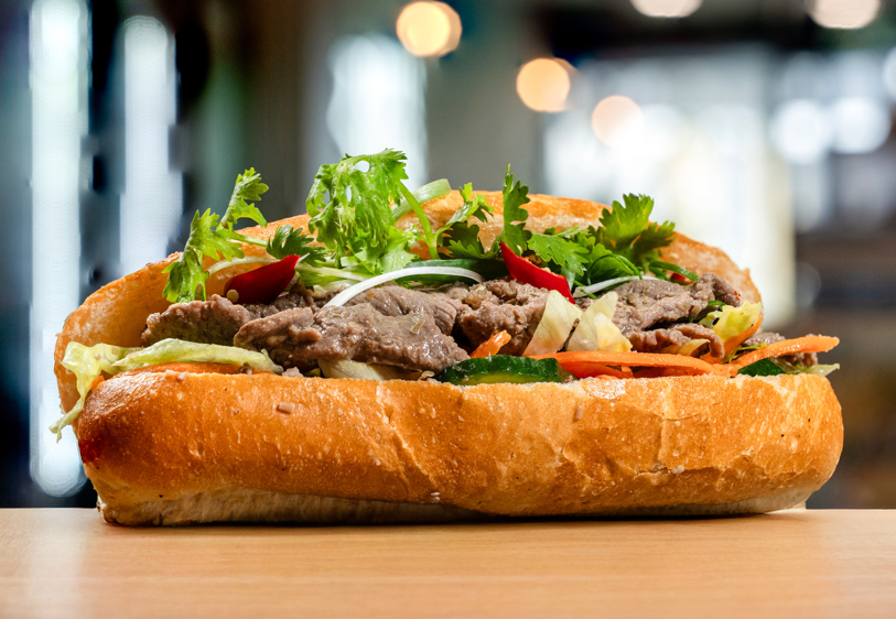 Philly cheesesteak sandwich with beef, lettuce, tomato, onions, and cilantro in a hoagie roll.