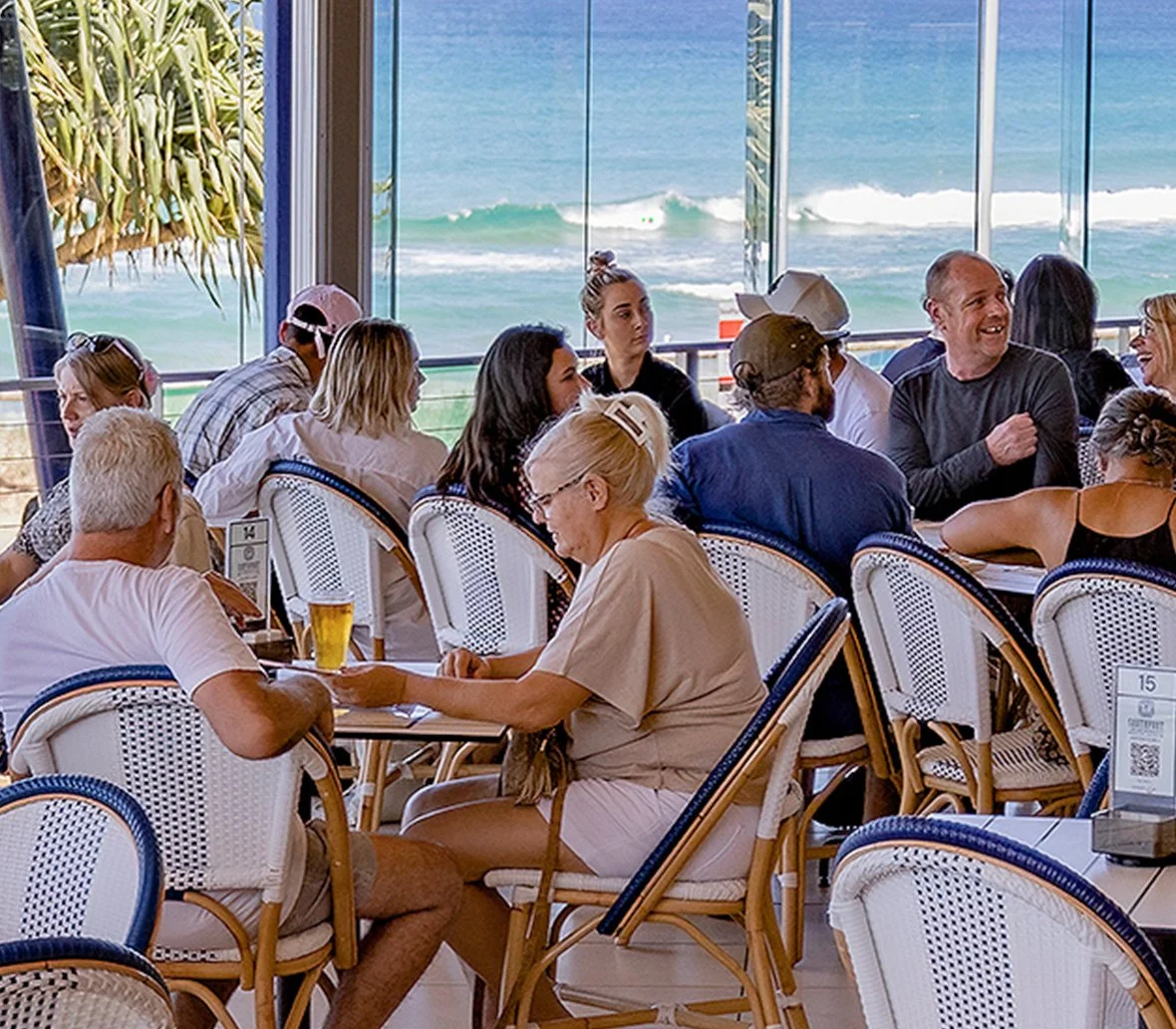 People dining at a restaurant with large windows overlooking the ocean, with waves visible outside.