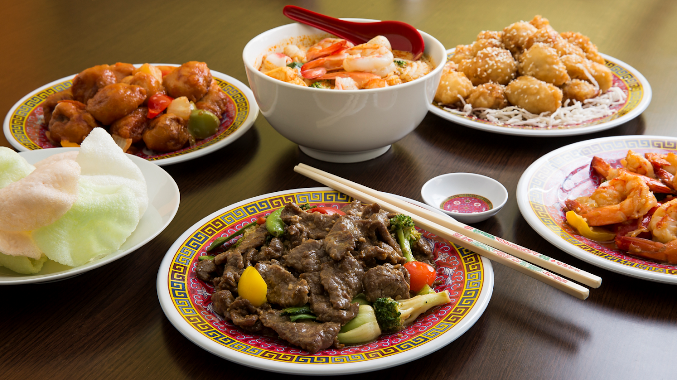 A variety of Chinese dishes on decorative plates, including orange chicken, beef with broccoli, shrimp in sauce, fried chicken, and scallion pancake, with a bowl of crab and shrimp soup in the center.