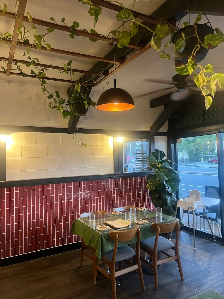Dining area with a small table set for four, surrounded by wooden chairs, in a restaurant with industrial decor, hanging plants, and a large window near the entrance.