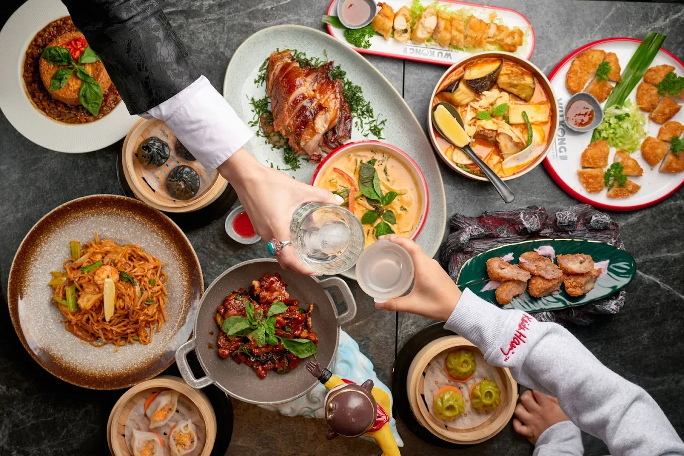 A variety of Asian dishes on a table, including noodles, dumplings, fried chicken, seafood, and various rice and vegetable dishes, with two people toasting with drinks.