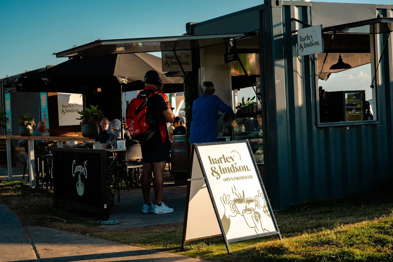 Outdoor coffee stand with a sign reading 'Harley & Hudson. Coffee. Open from 6 AM.' Several customers are ordering or waiting, with some seated at tables. The stand has a small, teal-colored structure with signs and plants, and is shaded by large umbrellas. The scene is illuminated by warm sunlight.
