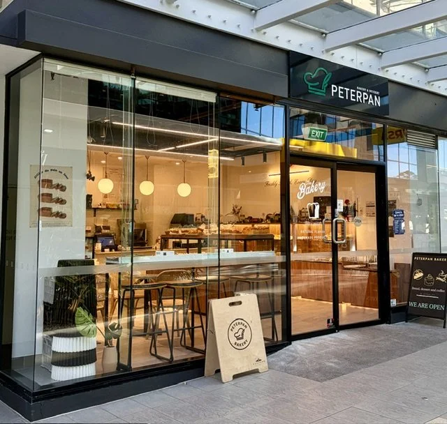 View of 'Peterpan Bakery & Dessert' in Southport with shiny glass facade illuminated by the gold lighting within and signage with white writing above the glass doors, some display boards out the front. yellow lighting