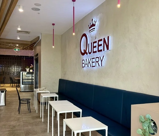 Inside 'Queen Bakery' in Queens Street Village located in Southport showing the modern new-fit out, a desert cabinet, comfortable looking padded seating with tables and an illuminated large logo on the wall crowned with a crown.