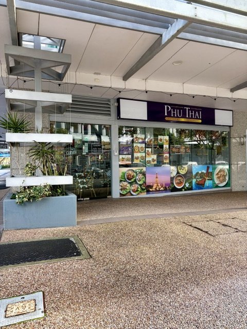 Outside of 'Phu Thai' Restaurant in Southport with light box signage on the wall in black, white and gold and bright pictures of Thai food on the windows with a planter box and greenery out the front.