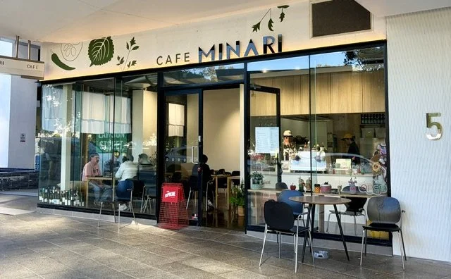 Street view of a Korean style Cafe called MINARI in Southport showing glass panel walls and front doors, table and chairs out the front and customers through the window seated within.