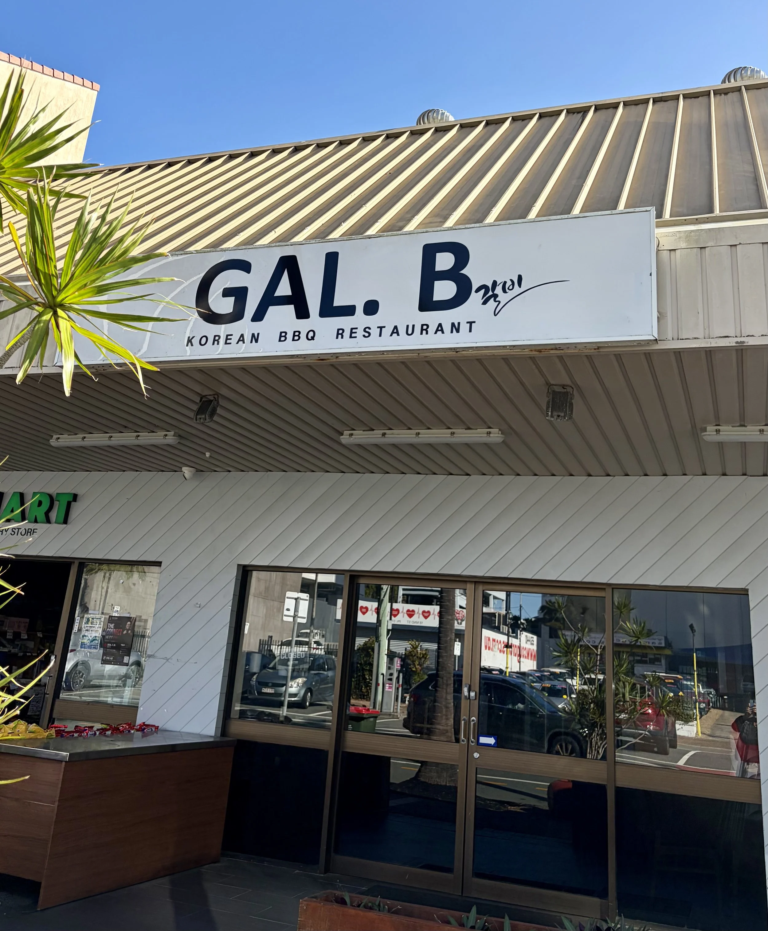 GAL. B Korean restaurant signage at front with black letters on white background with greenery in the image.