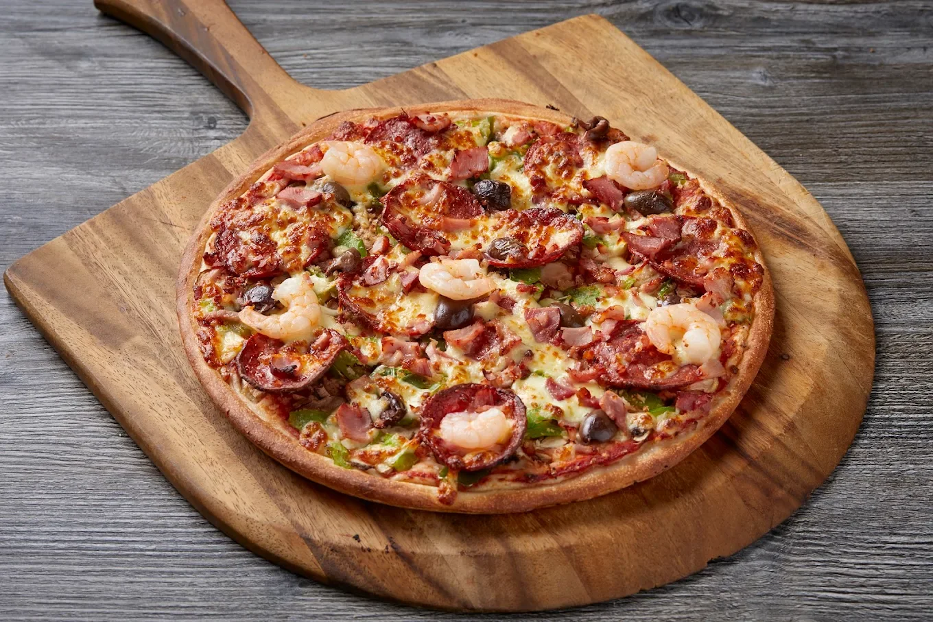 Close-up of a seafood pizza topped with shrimp, pepperoni, ham, green peppers, black olives, and melted cheese on a wooden pizza peel.