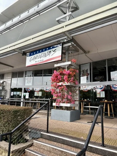Street view of 'Carolina's' Bar and Grill in Southport showing high glass facade, red white and blue signage, tables and chairs out the front and pink bougainvillea all around a column.