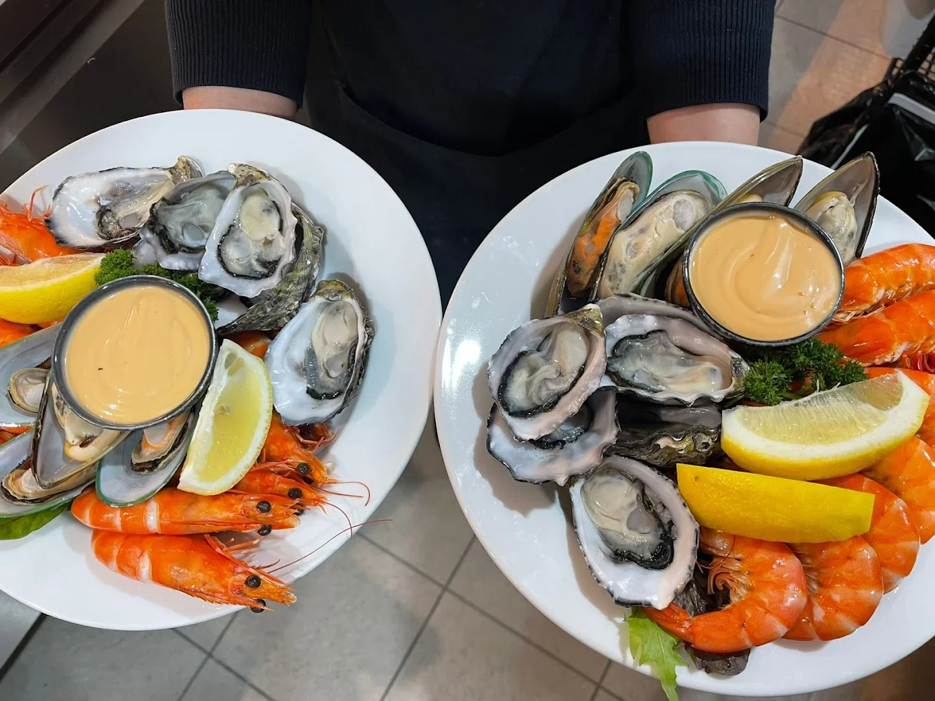 Two plates of seafood, including oysters on the half shell, cooked shrimp, lemon wedges, and a cup of creamy sauce.