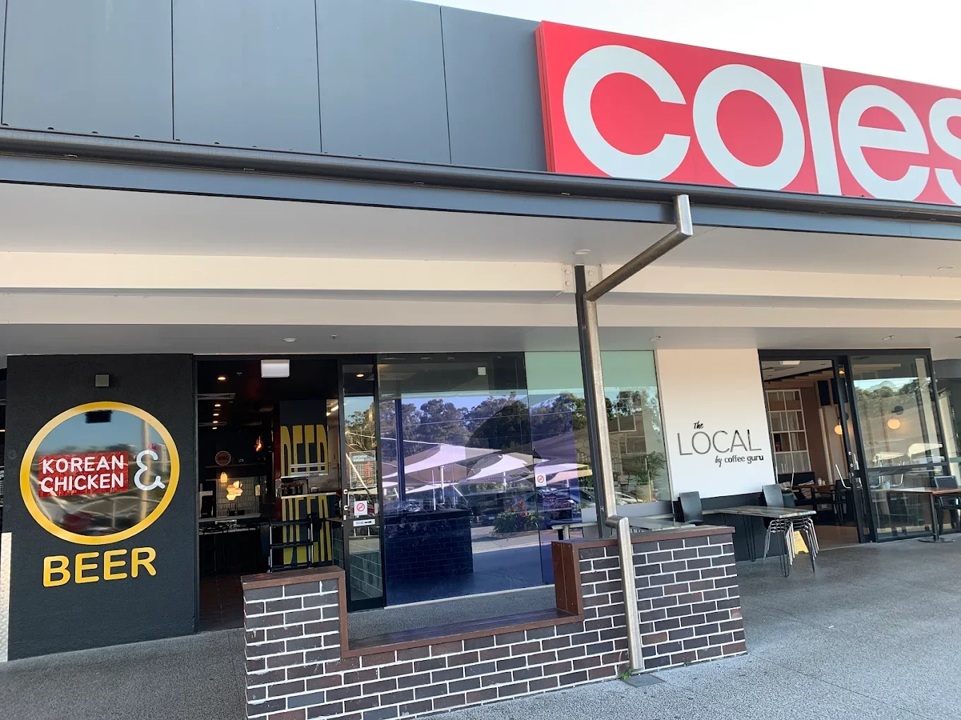Exterior view of a modern retail store with a large red and white sign that reads 'coles'. The storefront features glass doors and windows, with seating and umbrellas visible inside and outside. There is a sign indicating 'Korean Chicken & Beer' near the entrance.