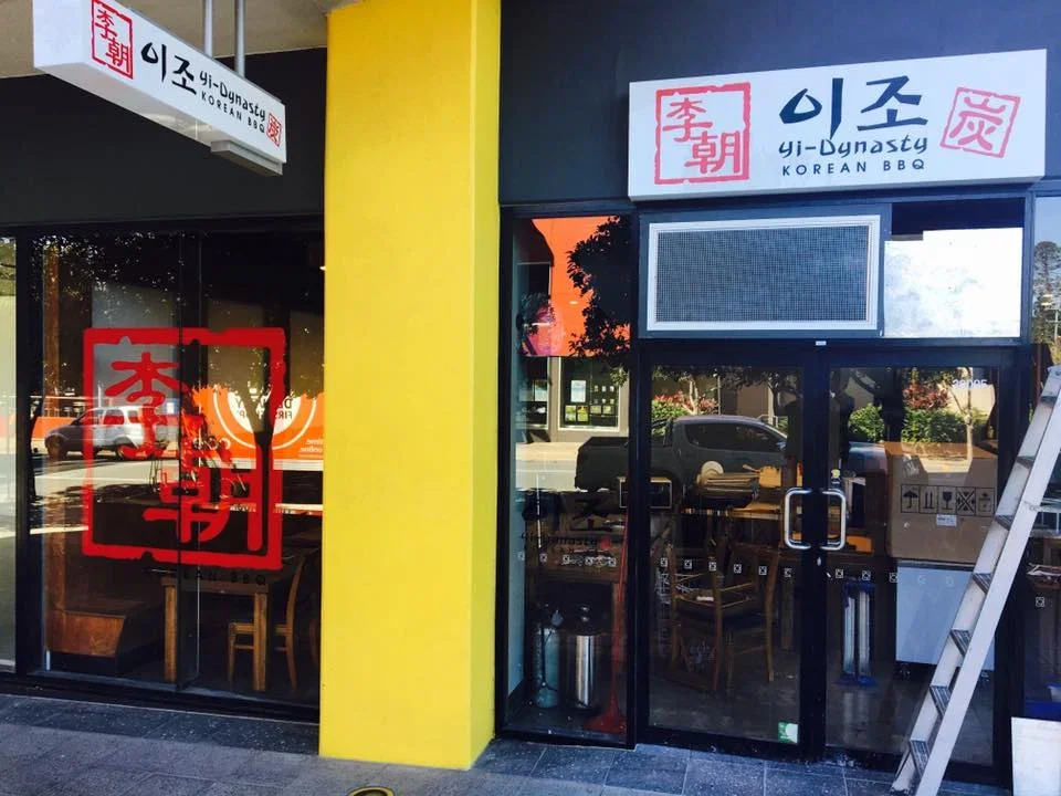 Exterior view of Yi Dynasty Korean BBQ restaurant with a glass door and window, featuring signage in Korean and English, and a red logo with Korean characters.