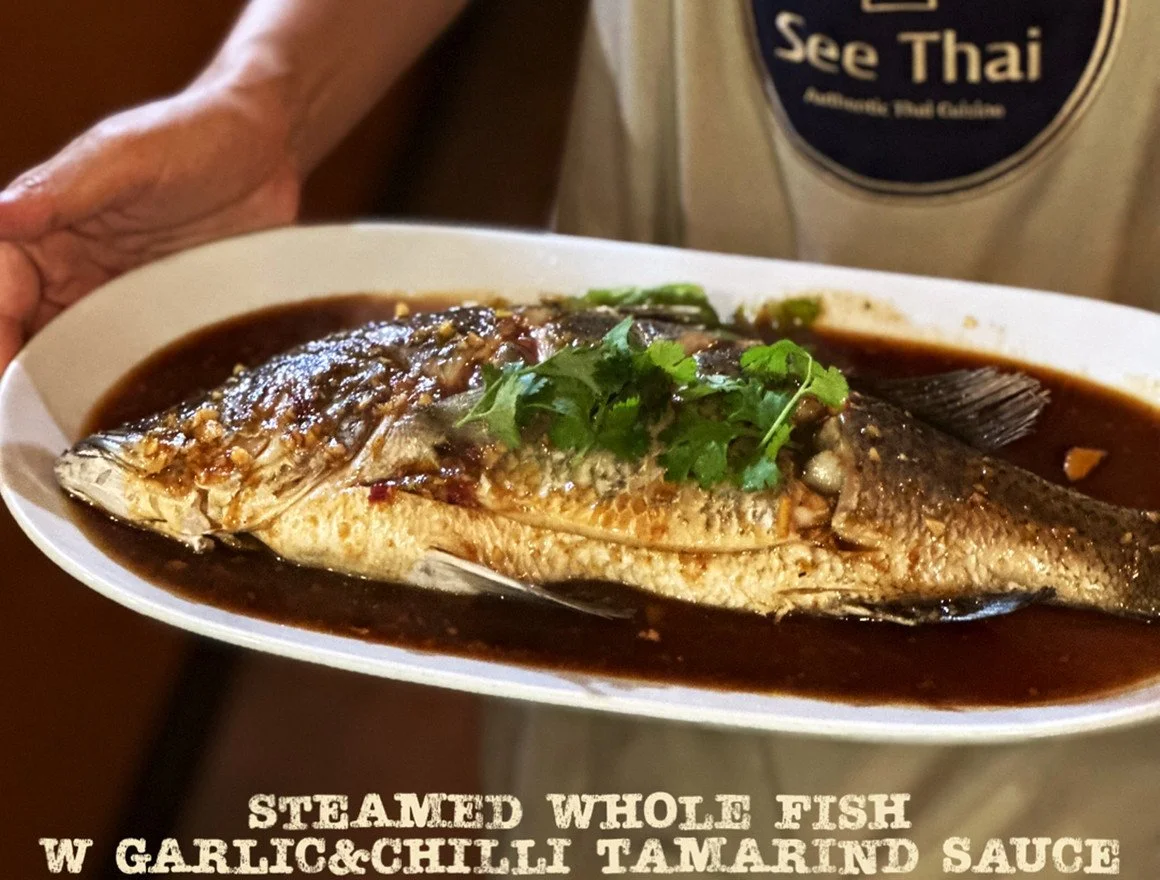 A whole steamed fish served on an oval white plate with garlic and chili tamarind sauce, garnished with a sprig of cilantro. The person holding the plate is wearing a shirt with a see Thai logo.