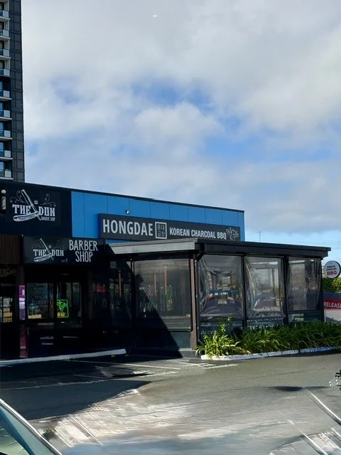 Street view of 'HONGDAE' Korean charcoal BBQ restaurant in Southport showing large black and white signage on bright blue building facade with outside covered eating area with plastic see-through sides for weather protection and some greenery.