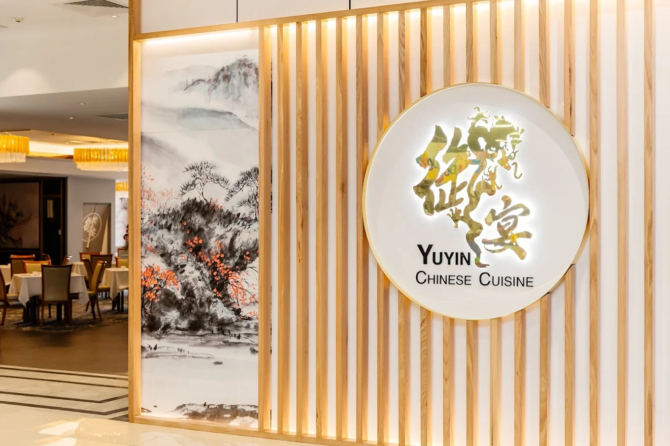 Interior view of a Chinese restaurant with a decorative wooden slat wall, a circular sign with Chinese characters and the words 'Yuyin Chinese Cuisine,' and traditional Chinese-style artwork on the wall.