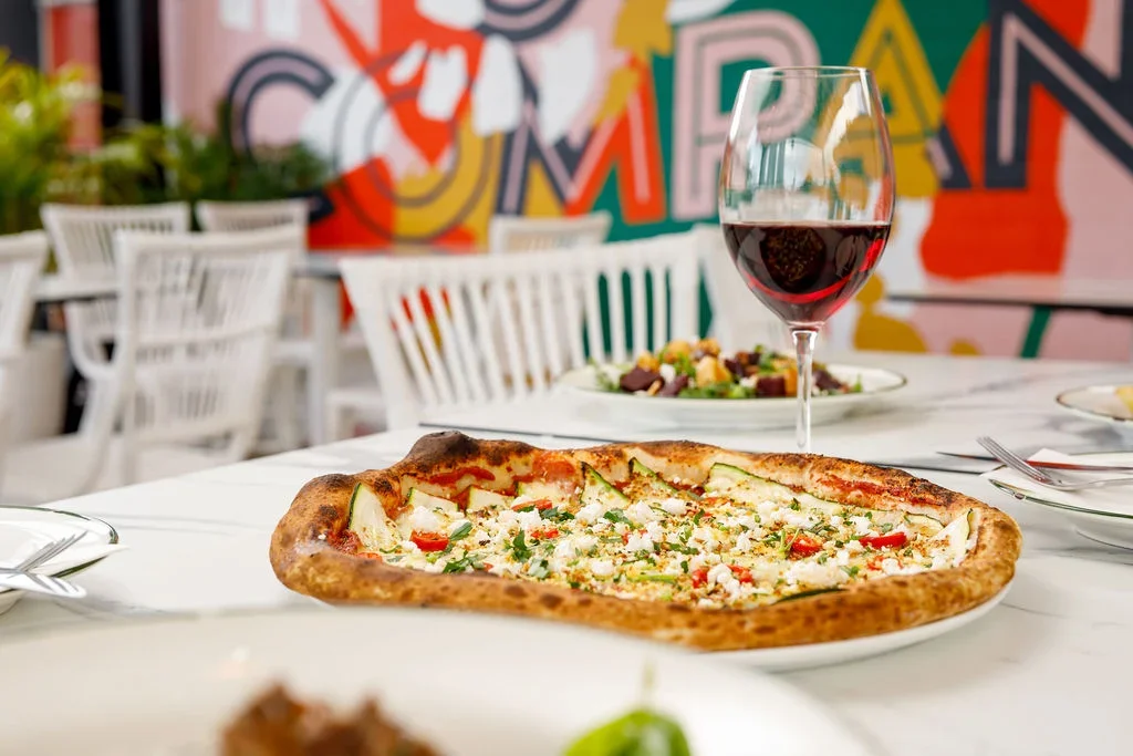A restaurant table with a rectangular pizza topped with cheese, vegetables, and herbs, a glass of red wine, and a plate of salad in the background. Bright, colorful mural on the wall.