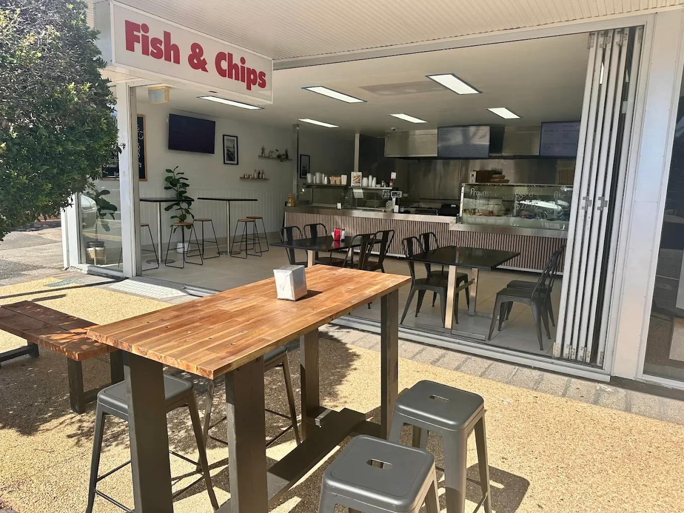 Small eatery with a large red and white 'Fish & Chips' sign. Outdoor seating with high-top tables and stools, no customers present. Indoor counter area visible, with a menu displayed on digital screens and a glass display case showing food items like prawns and oysters. Bright, minimal decor inside.