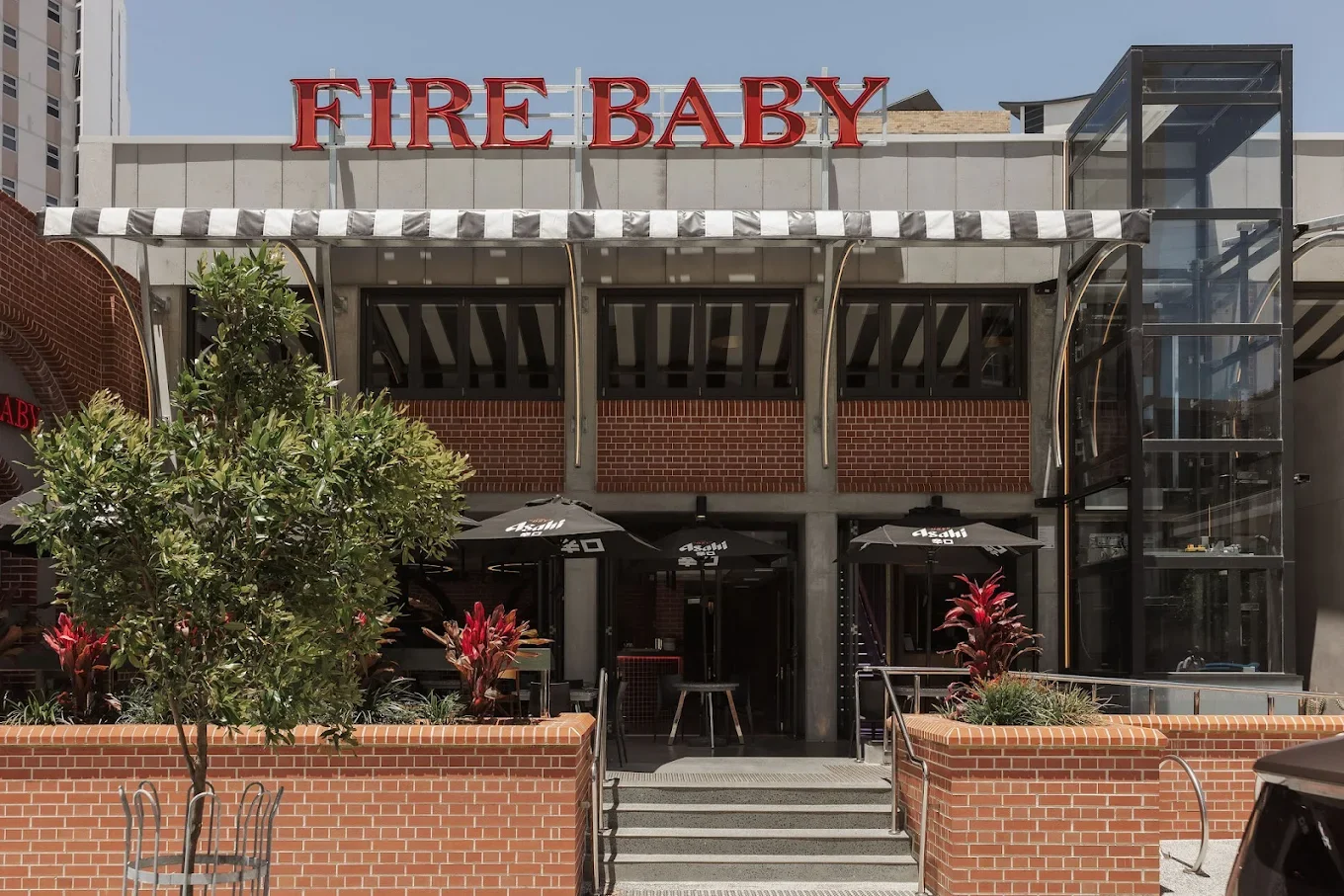 Exterior of a two-story restaurant named 'Fire Baby' with a brick facade, outdoor seating area with black umbrellas, plants, and a glass elevator on the right side.