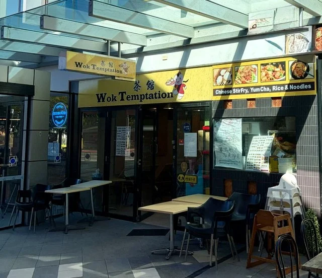 Front view of 'Wok Temptation' in Southport Central showing signage on bright yellow background, glass doors and tables and chairs.