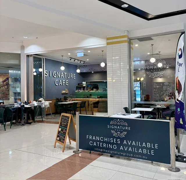 Inside a shopping mall, a cafe named Signature Cafe with tables, chairs, large signage on the inside wall against a dark teal background.
