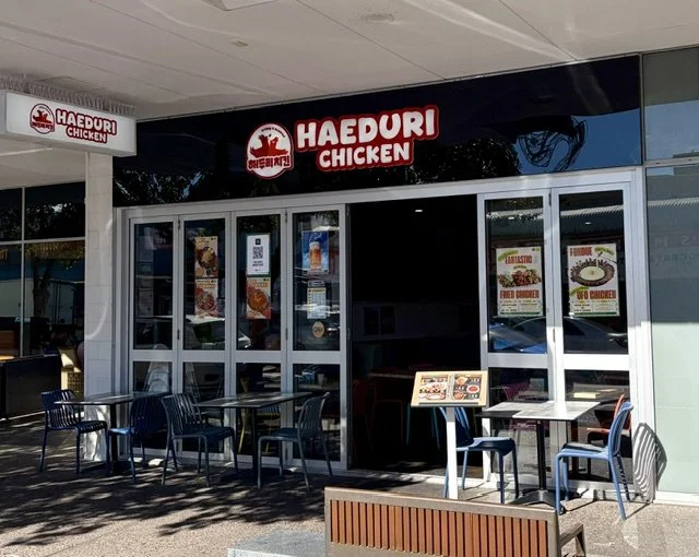 Exterior of a Korean restaurant named 'Haeduri Chicken' with tables and chairs out front, located in a shopping mall or complex