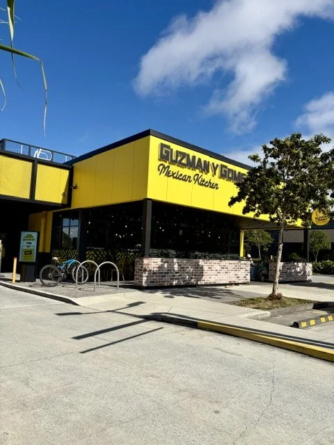 Bright yellow and black themed "Guzman Y Gomez' in Southport with a feature brick framed seating area out the front and there is also a bike in the bike racks on a sunny blue sky day.