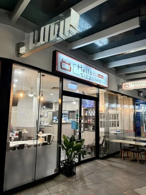 Night-time view from outside of Taiwanese style restaurant 'Hello Sizzling' in Southport showing glass frontage and doors, tables and chairs both inside and out and a large plant outside near the door.