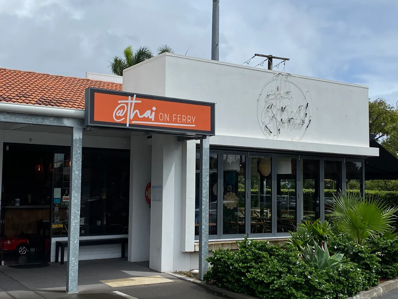 Outside view of a restaurant called '@thai on Ferry' with a modern white facade, large glass windows, and lush green plants in front.