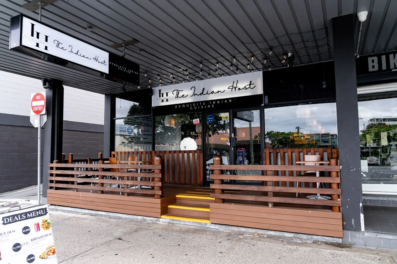 Exterior of The Indian Host restaurant with a wooden porch, signage, and a deals menu board outside.