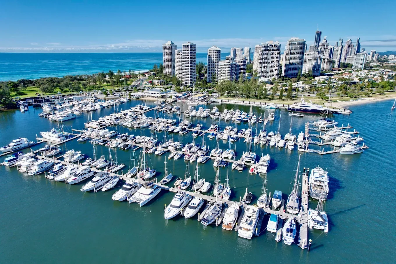 Aerial view of a marina with numerous boats docked, surrounded by a city skyline with high-rise buildings, green park areas, and the ocean in the background on a clear day.