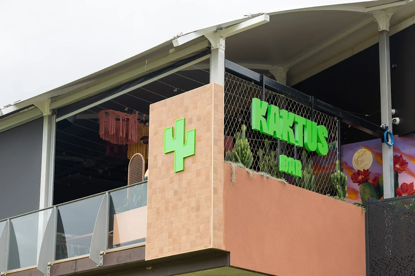 Exterior of a building with a green cactus logo and the words "Kaktus Bar" in bright green letters, with a balcony featuring potted cacti and colorful decorations.