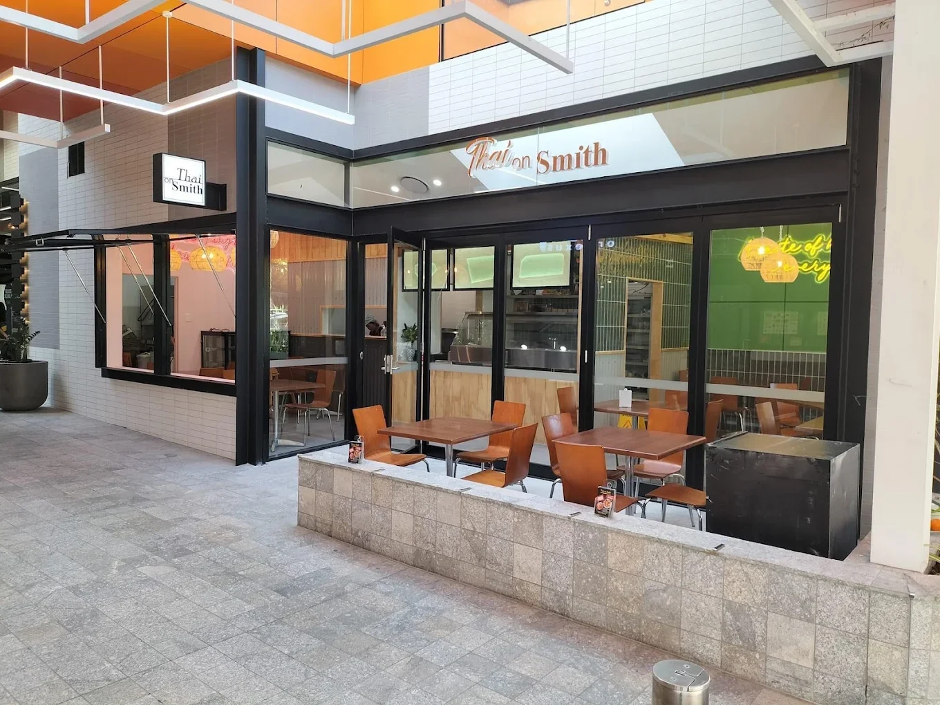 Exterior view of a modern restaurant with outdoor seating on a sidewalk, featuring wooden tables and chairs, large glass windows, and a white sign reading 'Thai on Smith'.