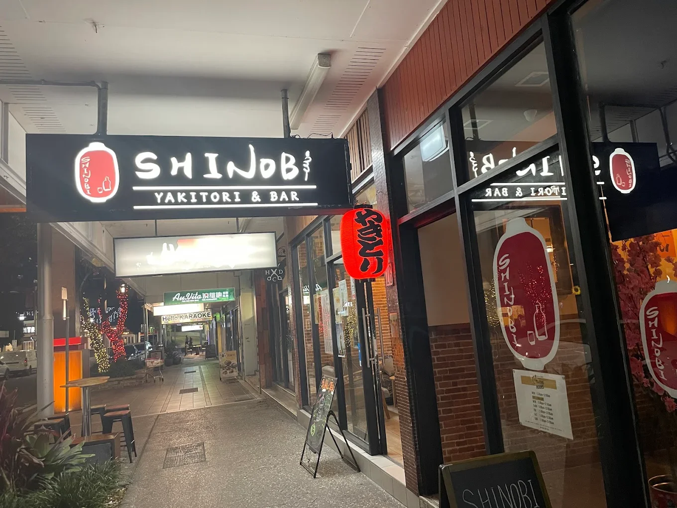 Night view of a restaurant storefront named 'SHINOBI Yakitori & Bar' with illuminated signage and red lanterns, located on a sidewalk.