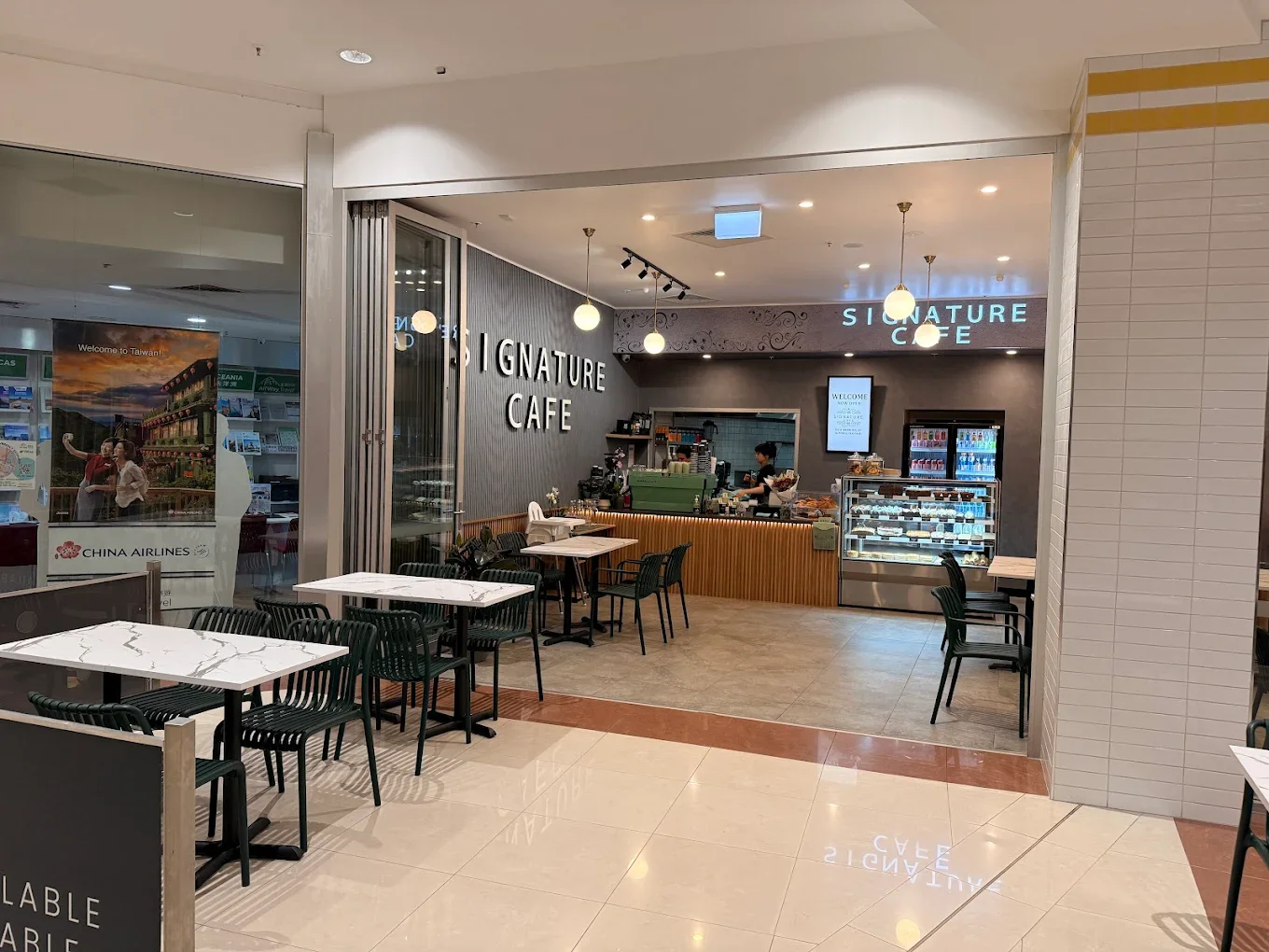 Inside a shopping mall, a cafe named Signature Cafe with tables, chairs, a counter, and a drink display case.