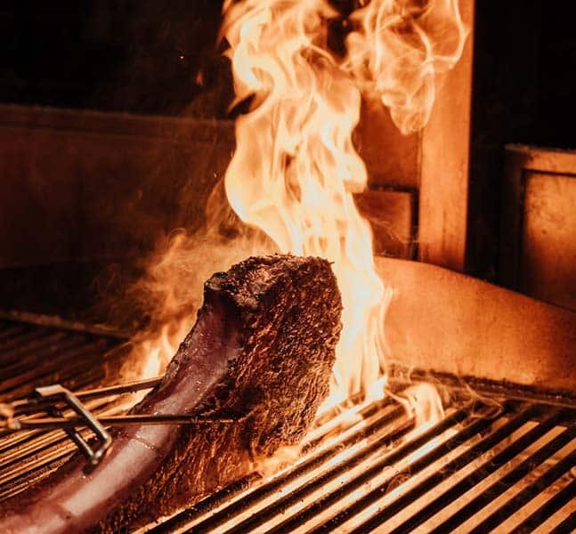 A cooked steak on a grill with flames and smoke surrounding it.