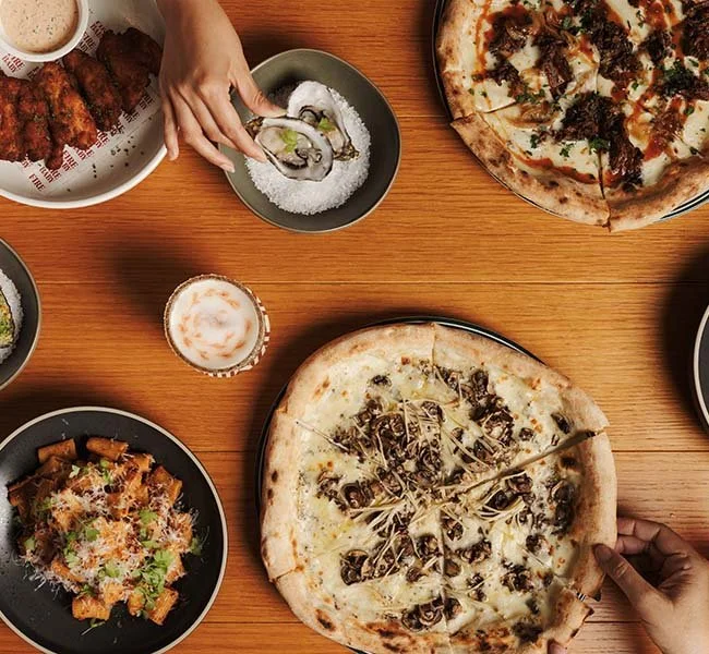 Various pizzas, a beer, and side dishes on a wooden table at a restaurant.