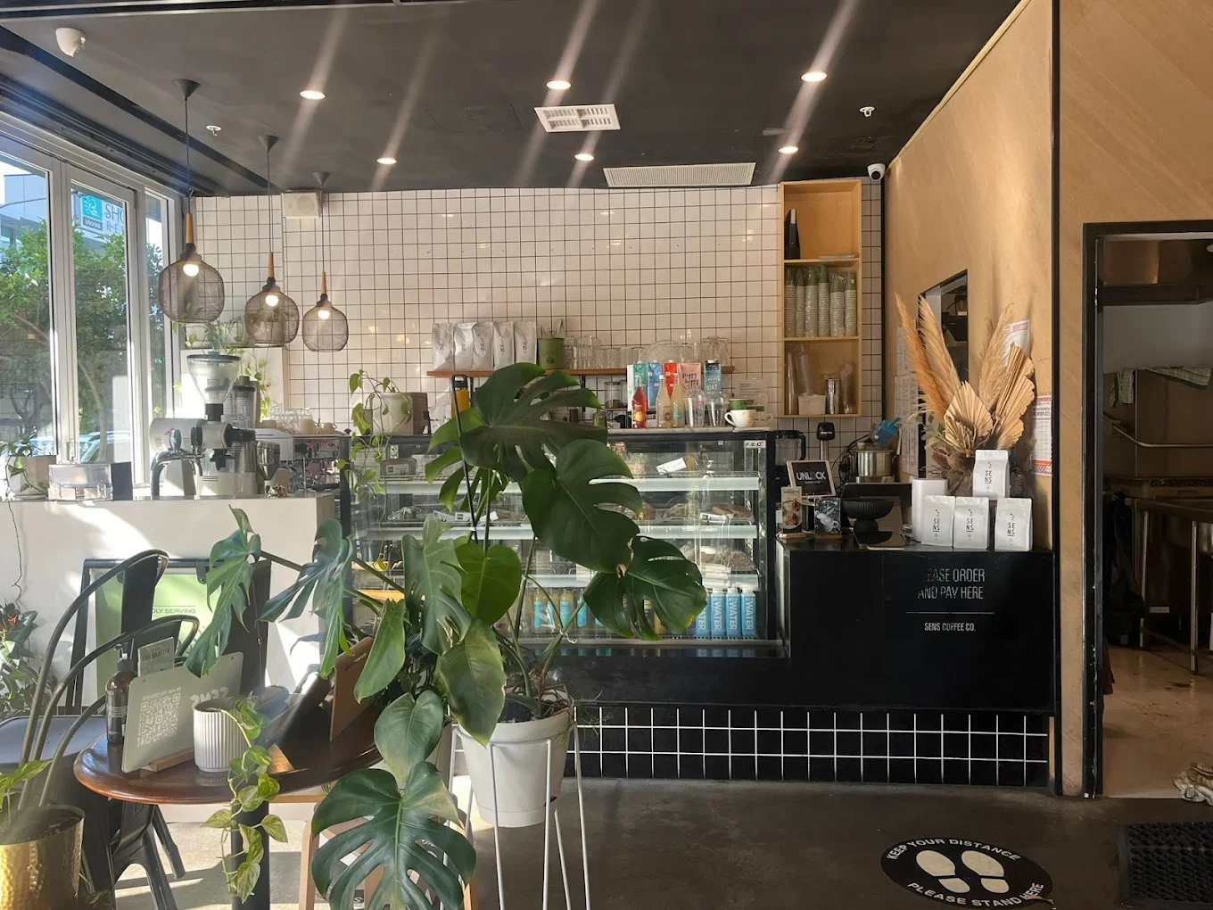 Interior of a modern coffee shop with a counter displaying baked goods, a white tiled wall, coffee equipment, hanging pendant lights, and green plants near large windows.