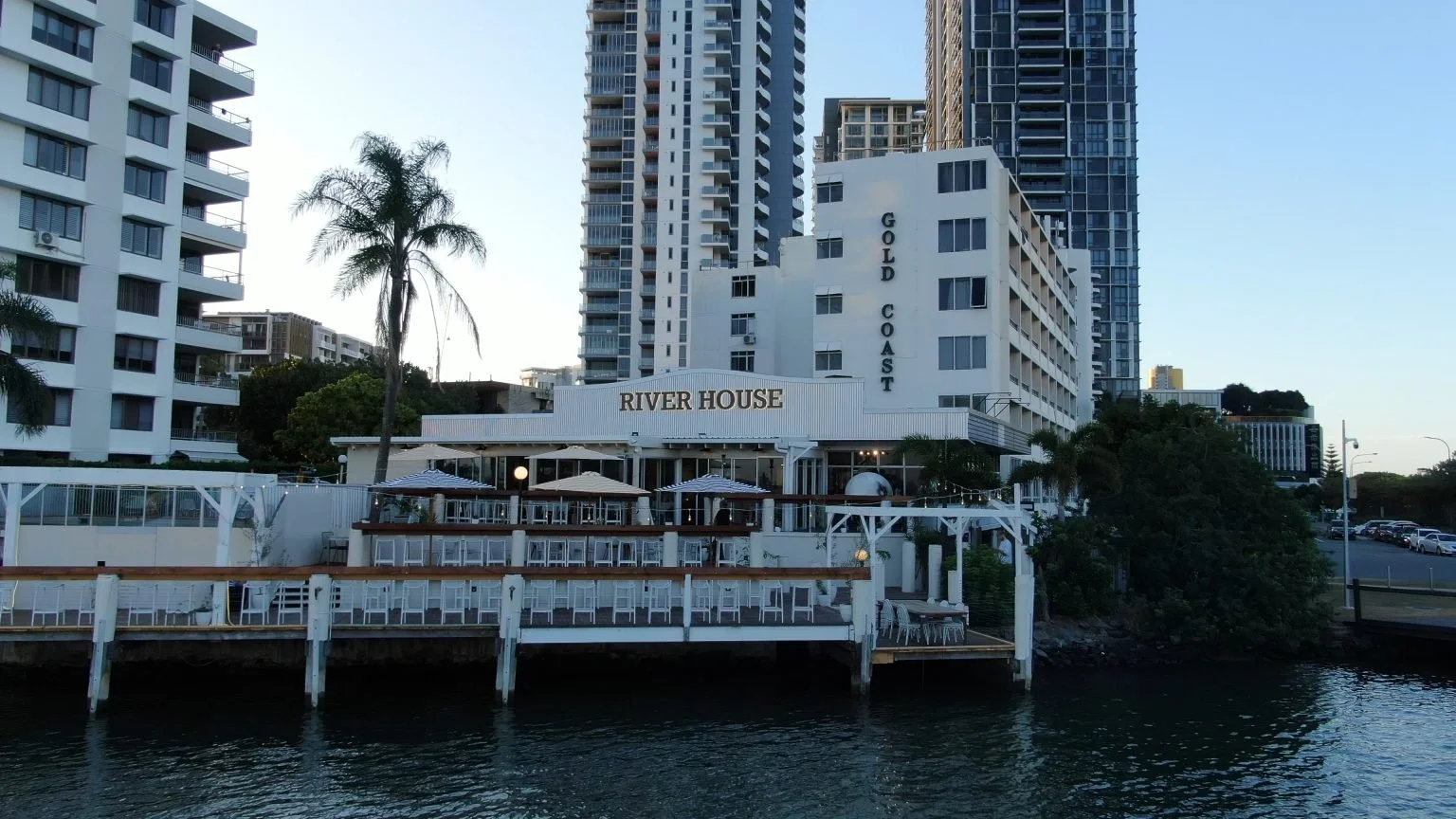 A waterfront building labeled 'River House' with outdoor seating and umbrellas, high-rise buildings in the background, a palm tree, and a parking lot on the right.