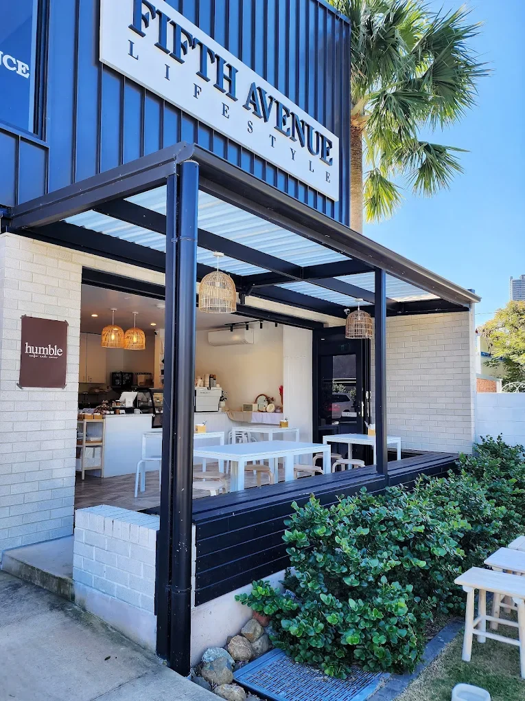 Exterior of a café with a sign that reads 'Fifth Avenue Lifestyle', outdoor seating with white tables and chairs, a small garden with green bushes, and a blue building façade with a palm tree nearby.