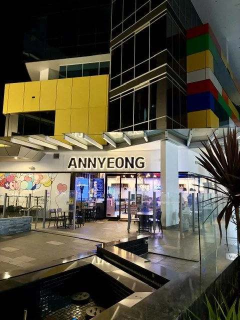 Night time view of Korean Restaurant 'ANNYEONG' in Southport showing the glassed outside seating area and feature garden and nib walls leading up to the restaurant. Large neon letters above feature the name.