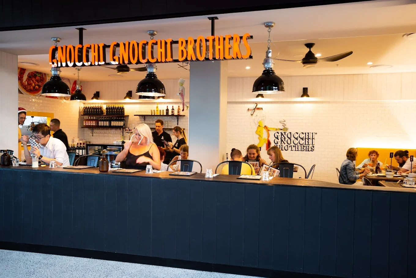 Inside Gnocchi Gnocchi Brothers restaurant with people dining at tables and a counter, restaurant sign hanging with orange letters, and a white brick wall with artwork.