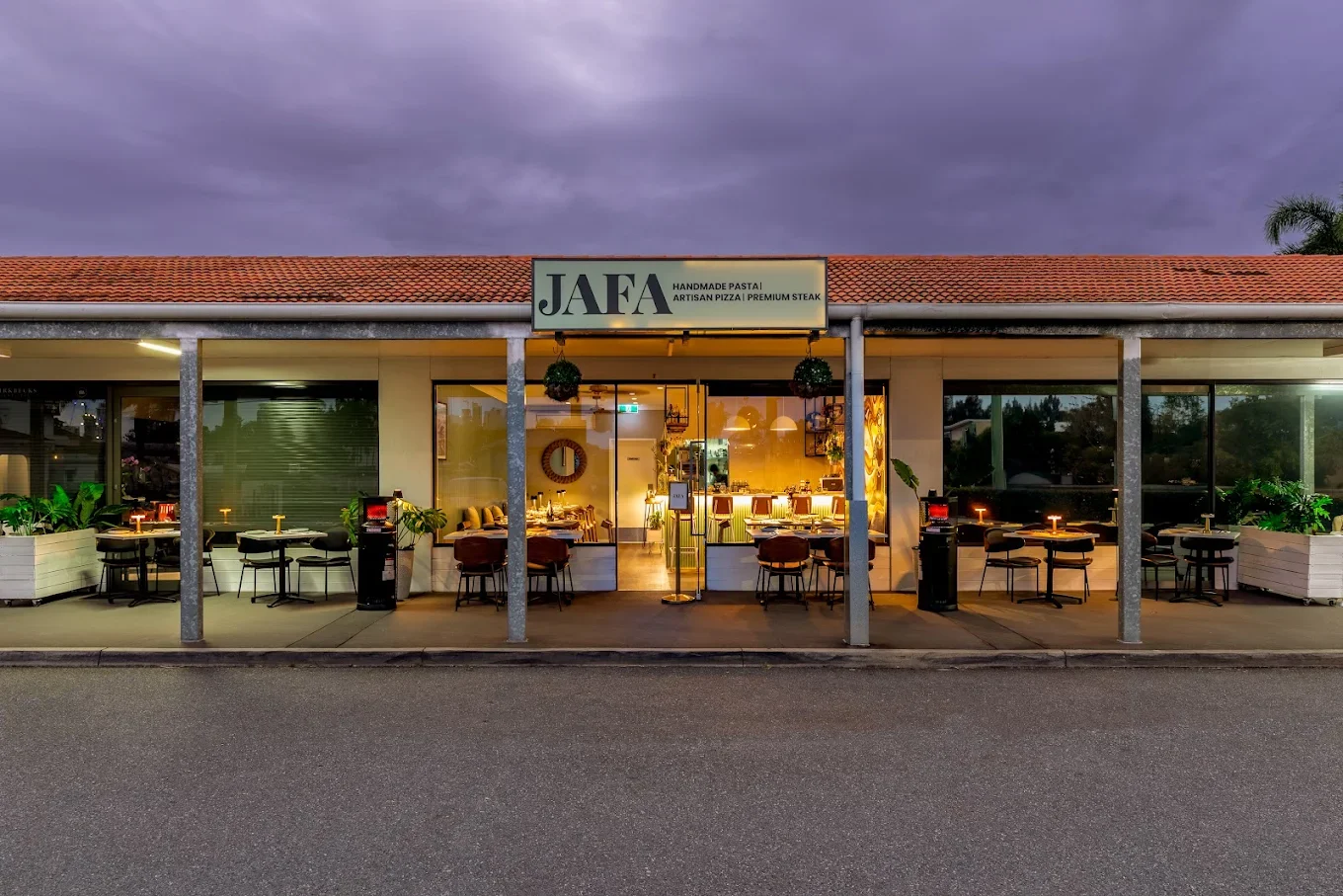 Exterior view of a restaurant called JAFA, with outdoor seating and large glass windows showing the interior, under a cloudy evening sky.